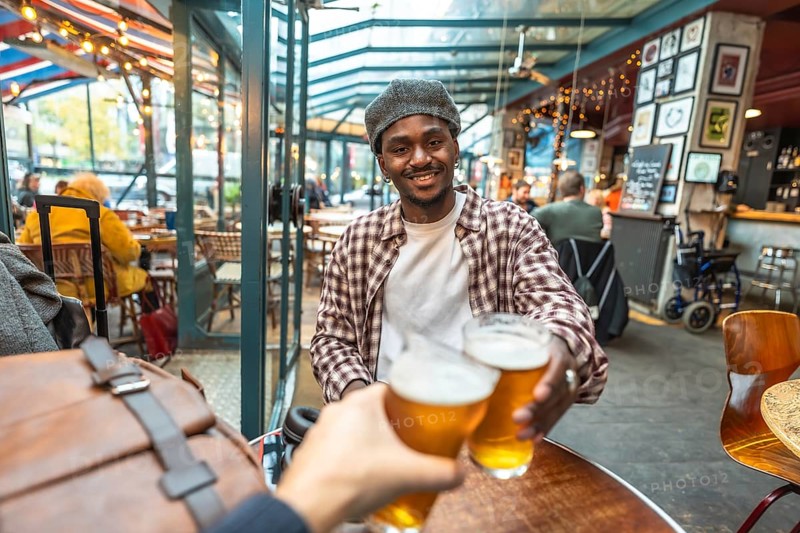 Young man smiling and clinking beer glasses with a friend in a casual pub, celebrating friendship, laughter and relaxed weekend vibes in an urban hangout