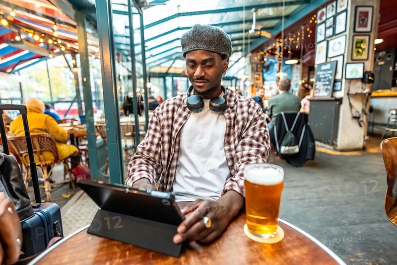 Young black man wearing headphones and flat cap working on digital tablet while sitting at a bar table with a glass of beer, embodying modern remote work and travel lifestyle