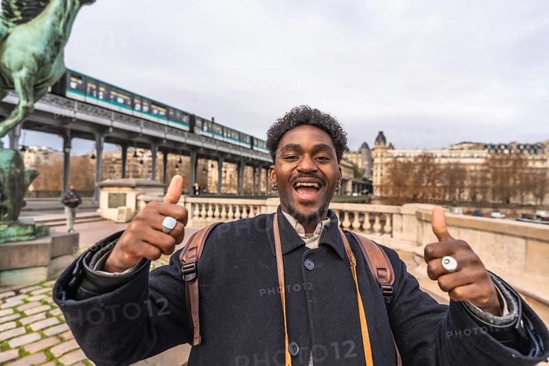 Enthusiastic young black man expressing happiness and positivity with a thumbs up gesture, traveling and exploring the city of paris with an elevated train in the background