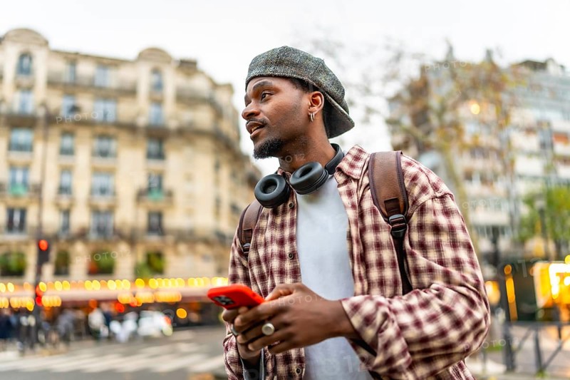 Young man using a smartphone and wearing headphones, a backpack, and a casual plaid shirt while exploring a city street with historic architecture in the background