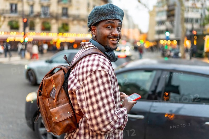 Young black man smiling, wearing a beret and backpack, holding a smartphone while walking on a busy city street with cars and buildings in the background