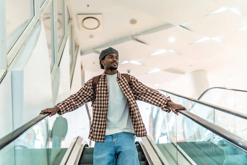 Young black man riding a modern escalator in a downtown shopping mall, wearing a backpack and stylish casual clothes, looking ahead while exploring urban retail spaces
