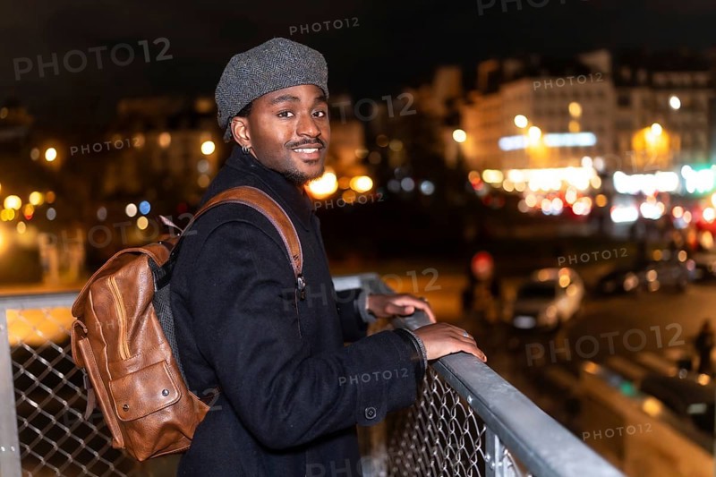 Young black man smiling while looking at the viewer, standing on a bridge overlooking the illuminated city of paris at night, enjoying urban exploration and a relaxed vacation