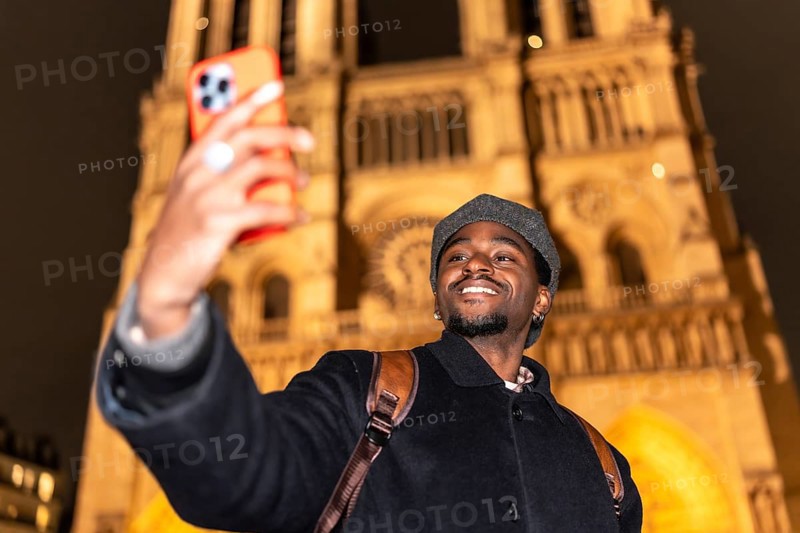Young man smiling and taking a selfie with a smartphone, capturing a memorable moment during a night cityscape tour of the iconic notre dame cathedral in paris, france
