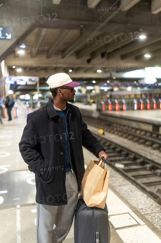 Young man with a baseball cap and glasses standing on a train station platform, holding a shopping bag and pulling luggage, looking out while waiting for his commute