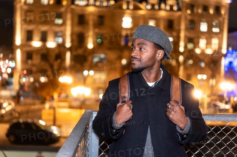 Young man in a beret and coat with backpack pauses on an illuminated paris street at night, gazing at city lights and reflecting on travel, solitude and urban discovery