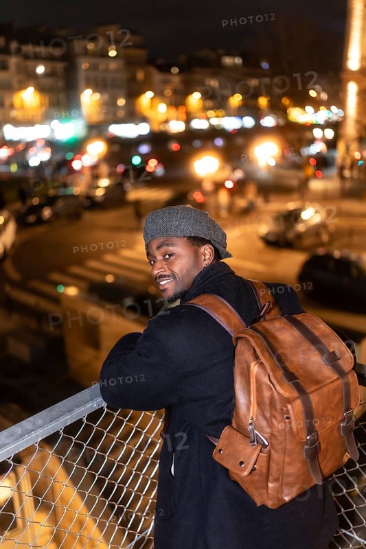 Young african man with a backpack and a cap. Smiling and looking at the camera. Standing on a bridge with blurred city lights and traffic visible in the background at night. Enjoying urban exploration