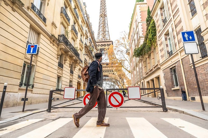 Young man with a backpack walking across a parisian crosswalk, historic buildings and the eiffel tower rising in the sunny autumn background, urban travel and sightseeing scene
