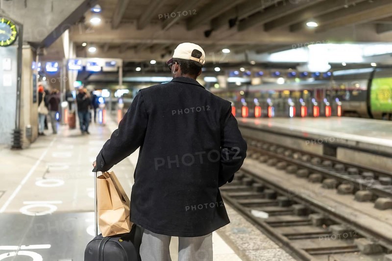 Man standing on a train station platform, seen from behind, pulling a suitcase and carrying a paper bag, waiting for public transport and contemplating his upcoming journey