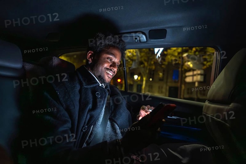 Young black man smiling and connecting with his smartphone, texting or browsing social media applications while traveling in the backseat of a car through the city at night