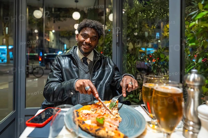 Young man smiling and cutting a delicious pizza slice with a knife and fork, enjoying a relaxing evening meal with a beer in a modern restaurant by the window