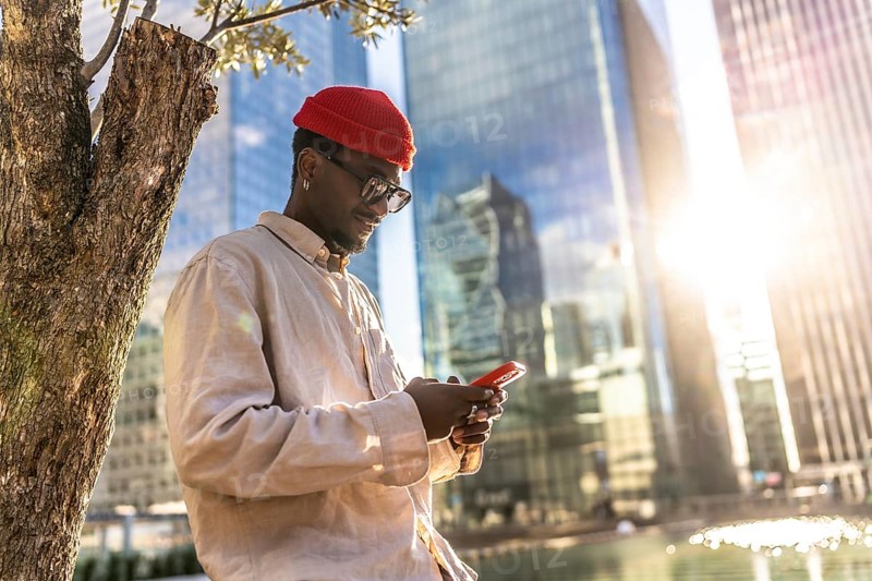 Young black man wearing a red beanie and sunglasses checking his mobile phone outdoors, standing by a tree in a modern urban environment with office buildings and sunlight