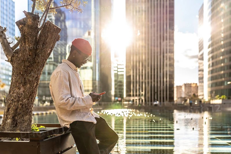 Young black man in casual attire concentrating on his smartphone while leaning by a tree, with modern skyscrapers and shimmering water in the bright evening sun