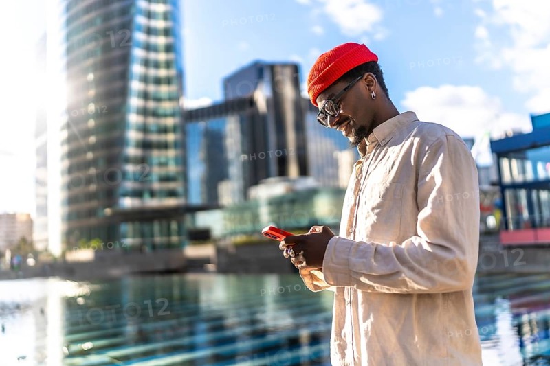 Young black man smiling while interacting with a smartphone outdoors, surrounded by modern city buildings and reflections, representing global communication and urban lifestyle