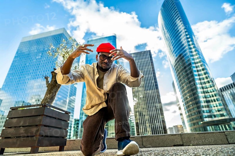 Young man wearing a red beanie and glasses, posing with hands raised in a low angle shot, expressing confidence and modern street style in a city environment with skyscrapers