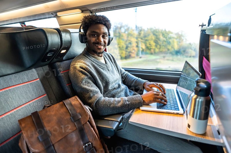 Young african man smiling, wearing headphones and typing on a laptop, working remotely or commuting on a business trip inside a modern train carriage next to a window