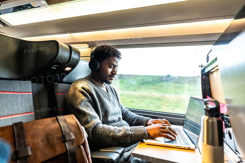 Young man wearing headphones concentrating on his laptop, working remotely or during a business trip while traveling by train, finding focus and productivity in motion