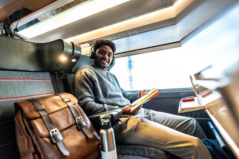 Happy black man wearing headphones and reading a book while traveling by train, sitting comfortably by the window enjoying the journey during a business trip