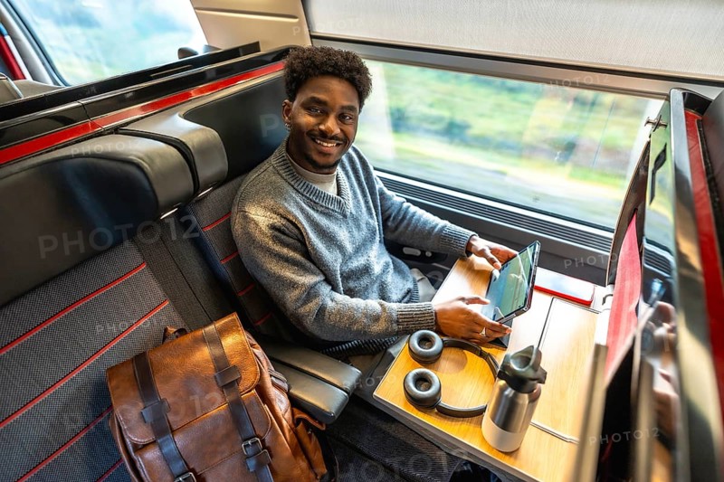 Young african american man traveling in a modern train, smiling and looking at the camera while working on his digital tablet, enjoying a comfortable and productive business trip