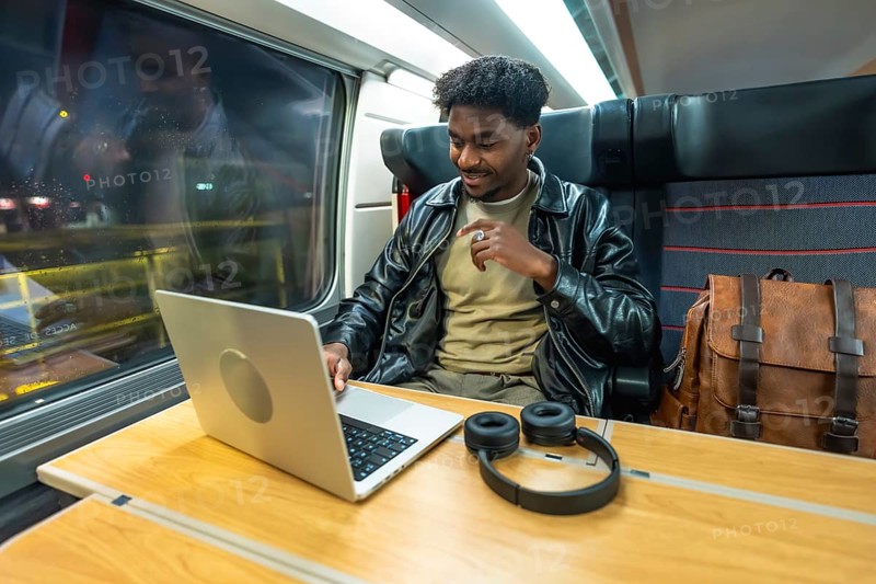 Young man using a laptop and headphones, smiling while traveling by train, reflecting modern business, remote work, and digital nomad lifestyle with connectivity on the go