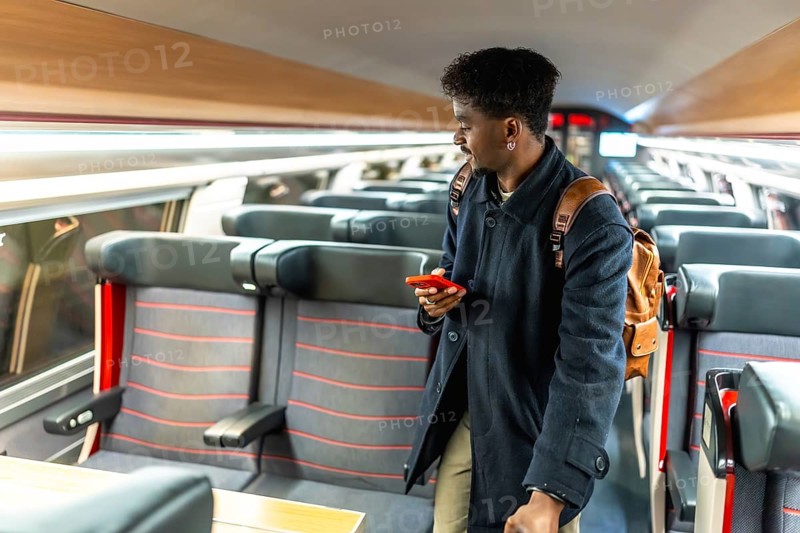 Young male passenger in a modern train carriage looking for his seat while checking travel information on a red smartphone during a business trip or commute