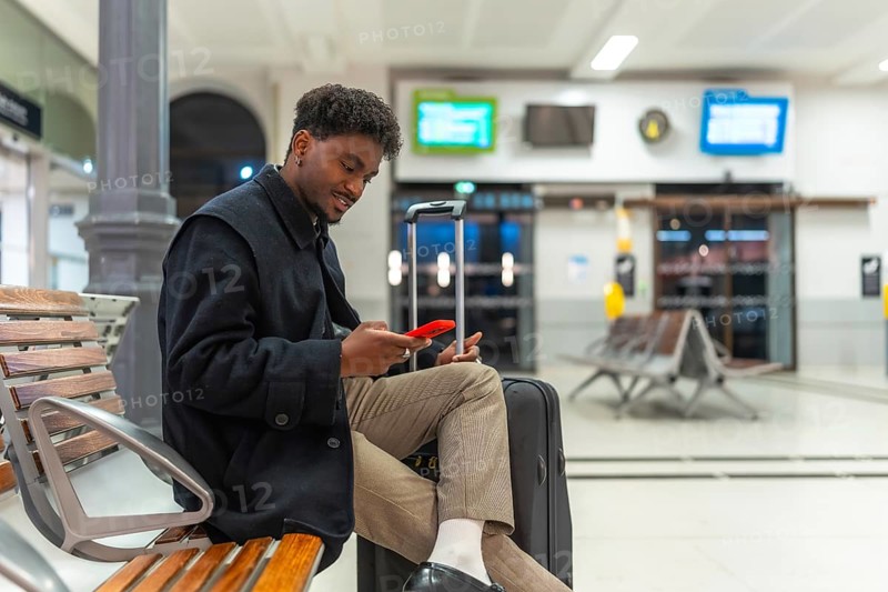 Young black man sitting comfortably on a wooden bench at a train station, using his red smartphone while waiting for transportation, with a black suitcase beside him