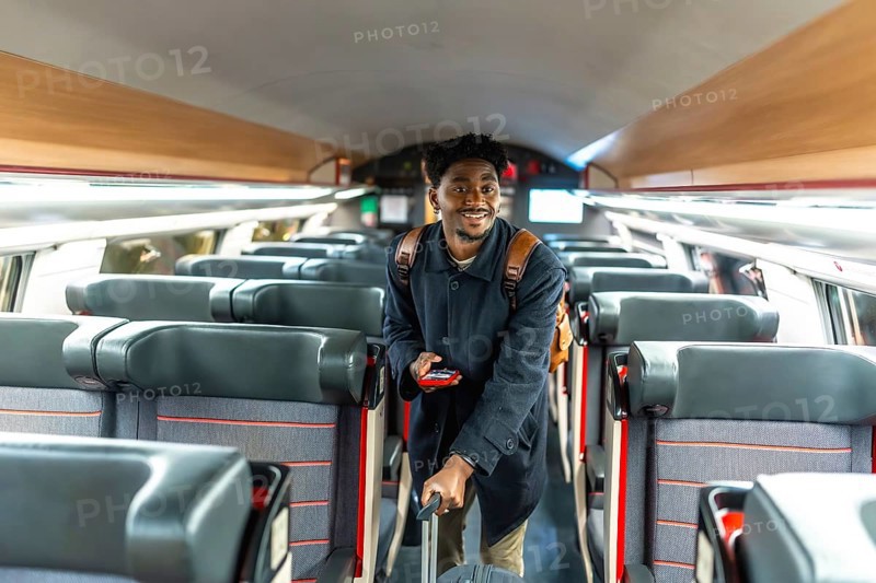 Young black man confidently walking through a train carriage, holding luggage and a mobile phone, representing modern travel, business journeys, and professional mobility