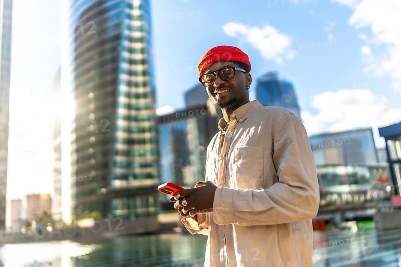 Young man smiling, holding and looking at a mobile phone, standing in a vibrant urban environment with sun drenched modern glass skyscrapers reflecting the sky