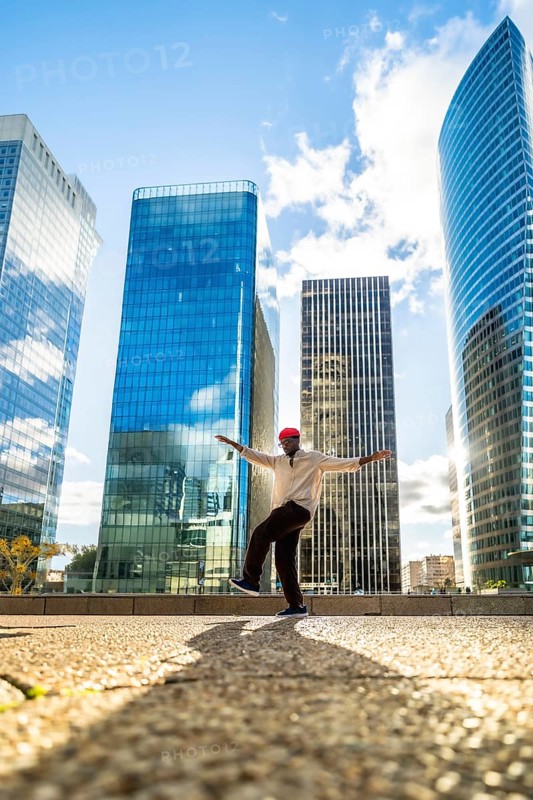 Stylish black man balancing on one leg with outstretched arms, expressing joy and freedom against a backdrop of modern skyscrapers in a sunny urban district