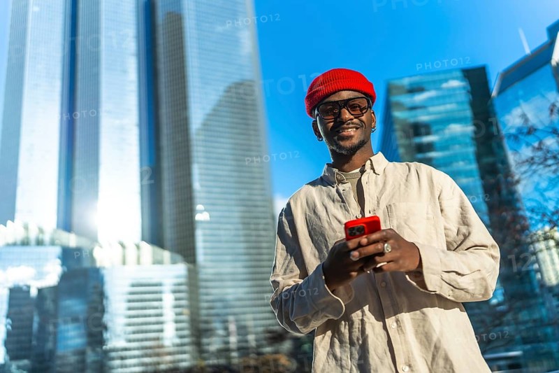 Young man smiling and checking content on his smartphone, linking to technology and modern communication while standing in a business district with glass skyscrapers under a blue sky