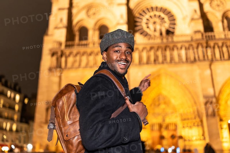 Joyful african american man with a backpack smiling and pointing, experiencing a nighttime tour of notre dame cathedral during his european travel adventure in paris, france