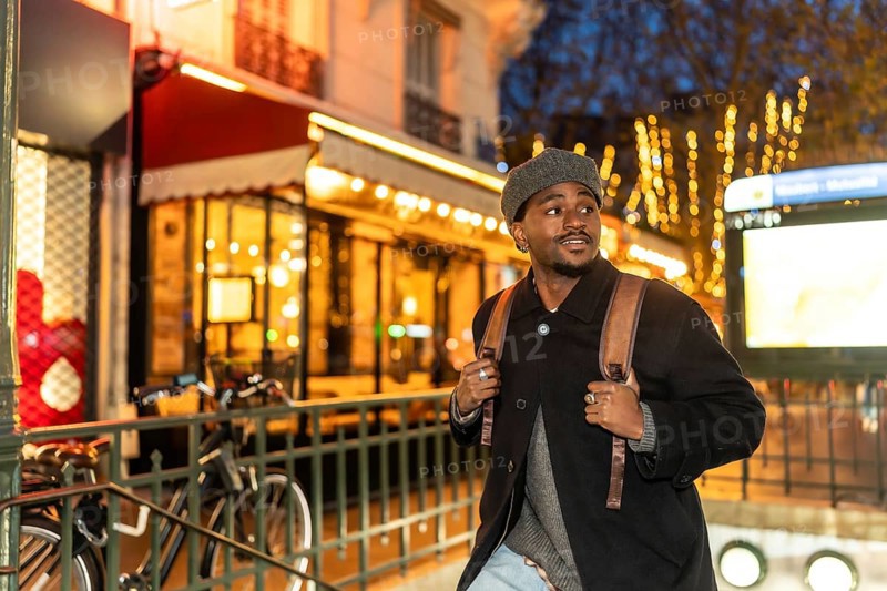 Young black man with a backpack exploring the illuminated streets of paris at night, enjoying an urban adventure near a subway entrance and a typical french cafe