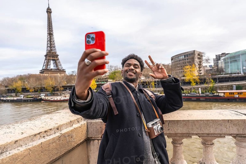 Young man smiling and making a peace sign while taking a selfie with a smartphone, capturing his happy travel experience with the iconic eiffel tower and seine river in the background