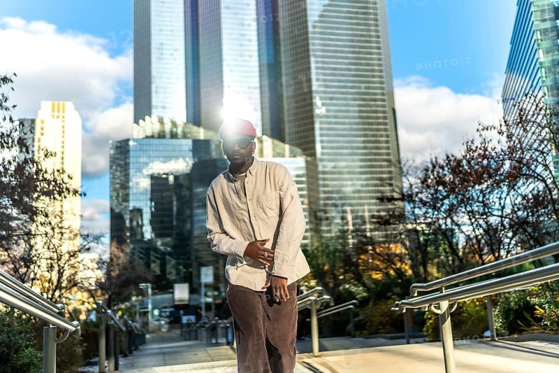 Young man wearing casual street style clothes and sunglasses, looking at camera while posing on steps with modern skyscrapers reflecting bright sunlight in a vibrant urban environment