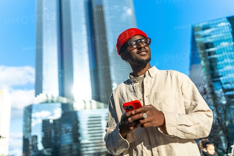 Young black man wearing a red beanie and glasses, smiling and looking up while holding a red smartphone with modern skyscrapers in the urban background