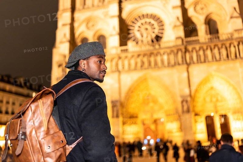 Young black man with a backpack and flat cap looking out while walking in front of notre dame cathedral at night, exploring the culture and architecture of paris