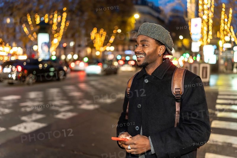 Smiling young black man wearing a hat and backpack, holding a smartphone while looking away, standing on a crosswalk in a busy city street illuminated by festive lights at night