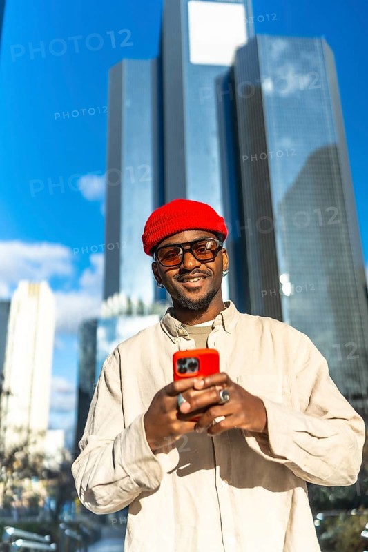 Stylish young black man wearing a red beanie and sunglasses, holding a bright red smartphone and smiling confidently in a modern city environment with skyscrapers under a clear blue sky