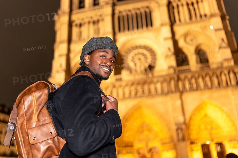 Young black man smiling, discovering french culture and history, standing in front of the illuminated notre dame cathedral in paris, celebrating travel and tourism