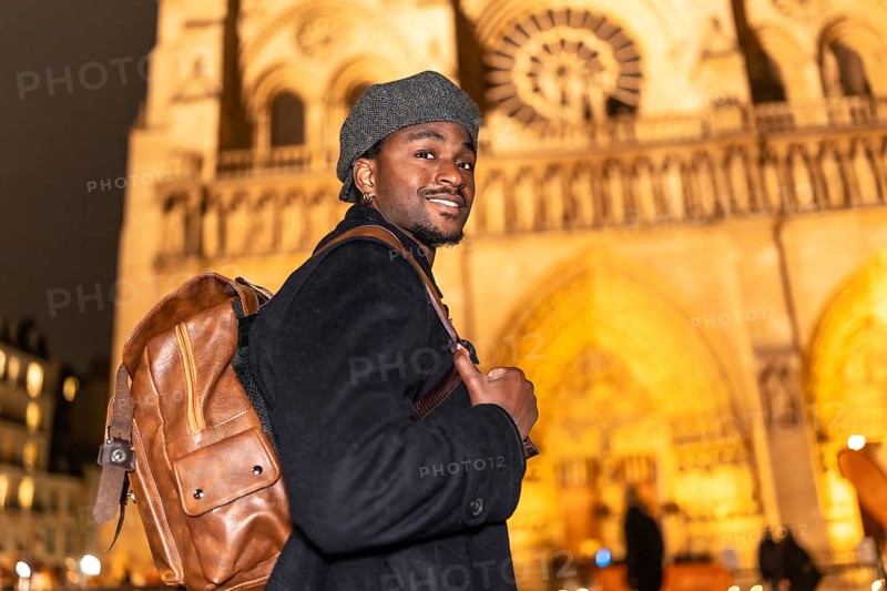 Young black man smiling while wearing a beret and backpack, standing in front of the illuminated notre dame cathedral in paris france at night, enjoying global tourism