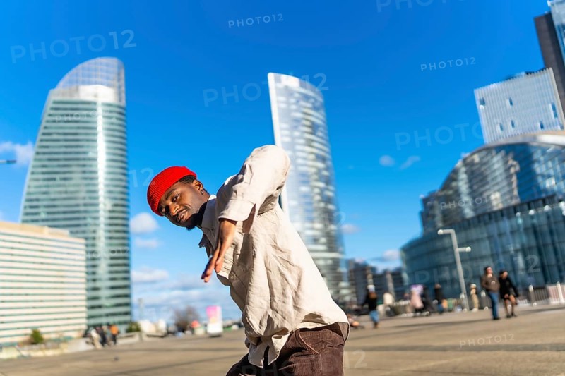 Young black man performing dynamic street dance in la defense, paris, striking a confident pose against sunlit contemporary skyscrapers and a bright blue sky