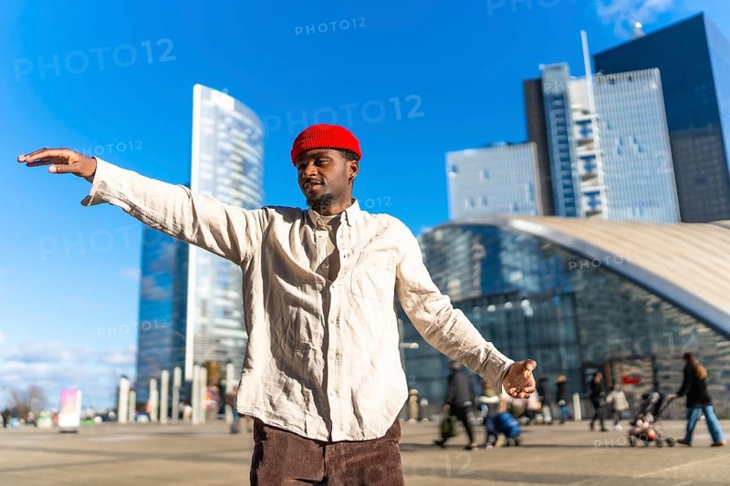Black man wearing a red beanie and light shirt expressing creativity through dancing, arms spread wide outdoors in a modern cityscape with skyscrapers