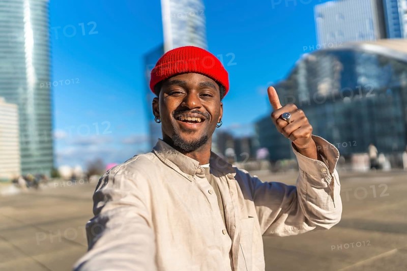 Young black man with a red beanie taking a close up selfie in an urban setting, smiling and giving a thumbs up gesture for success and approval on a sunny day