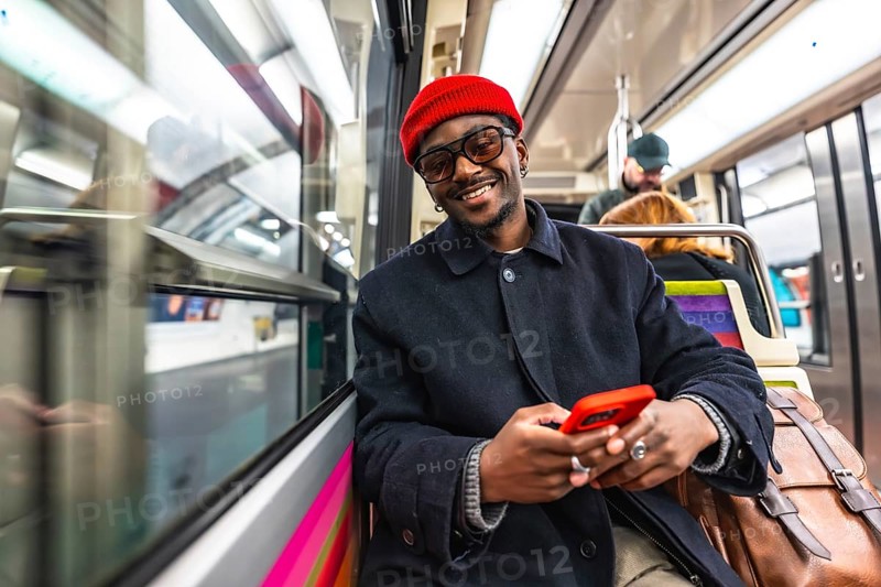 Young man smiling directly at the camera while holding and using a red smartphone, sitting on a subway train during his daily commute and journey through the city