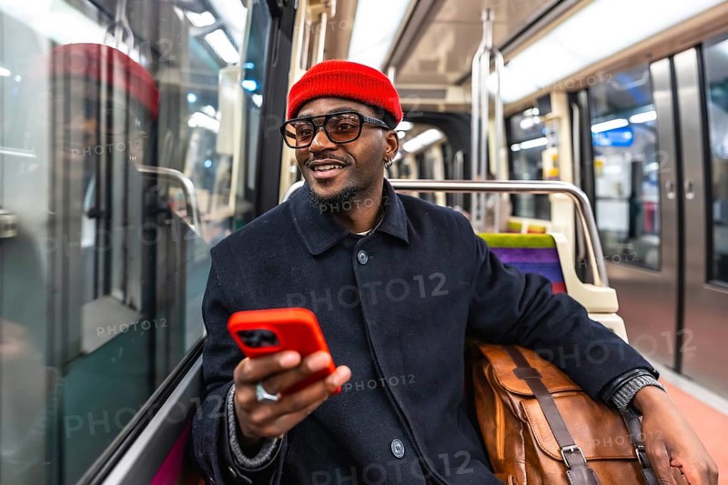 Young smiling african american man in a stylish coat and red beanie sits on a city train, holding a smartphone and browsing apps during a winter commute
