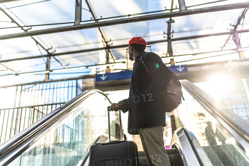 African american man traveling with a suitcase and backpack, riding an escalator upward in a modern train station or airport terminal with bright sunlight