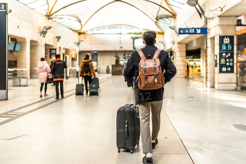 Businessman walking through a modern train station, pulling a suitcase and wearing a backpack, highlighting concepts of travel, commuting, business trips, and transportation