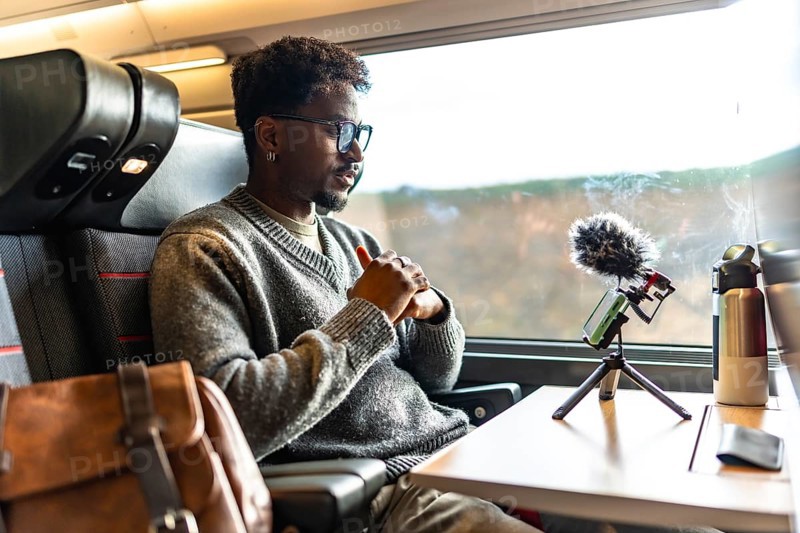 Young black man vlogging on a train, using smartphone and microphone on a tripod during a business trip remote work, focused content creator and digital nomad on the move