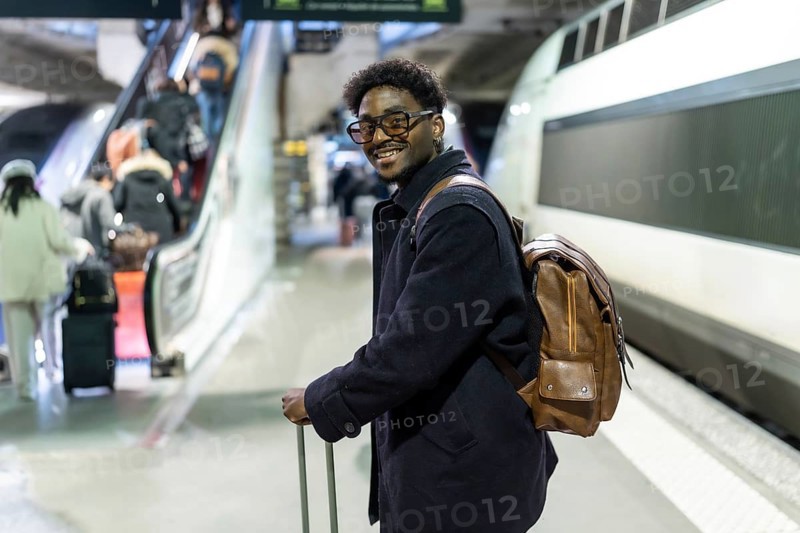 Smiling black man stands on a modern train platform with suitcase and backpack, ready for a business trip or commute confident, stylish, and prepared for urban travel and work