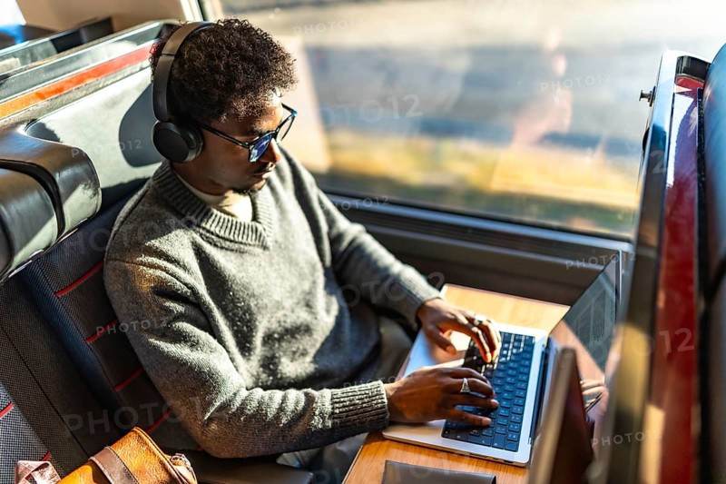 Young black man focused on laptop on a train, wearing headphones and glasses while working remotely on a business trip, exemplifying digital nomad mobility and mobile office productivity